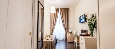 A hotel room with a bed, a desk, a chair, a small table, a mirror, a TV, and a vase with flowers.