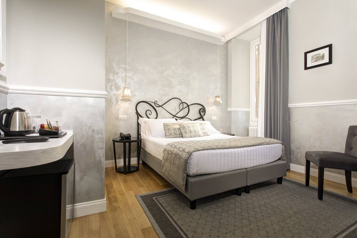 A modern hotel room with a bed, bedside table, and a desk with a kettle and coffee maker.