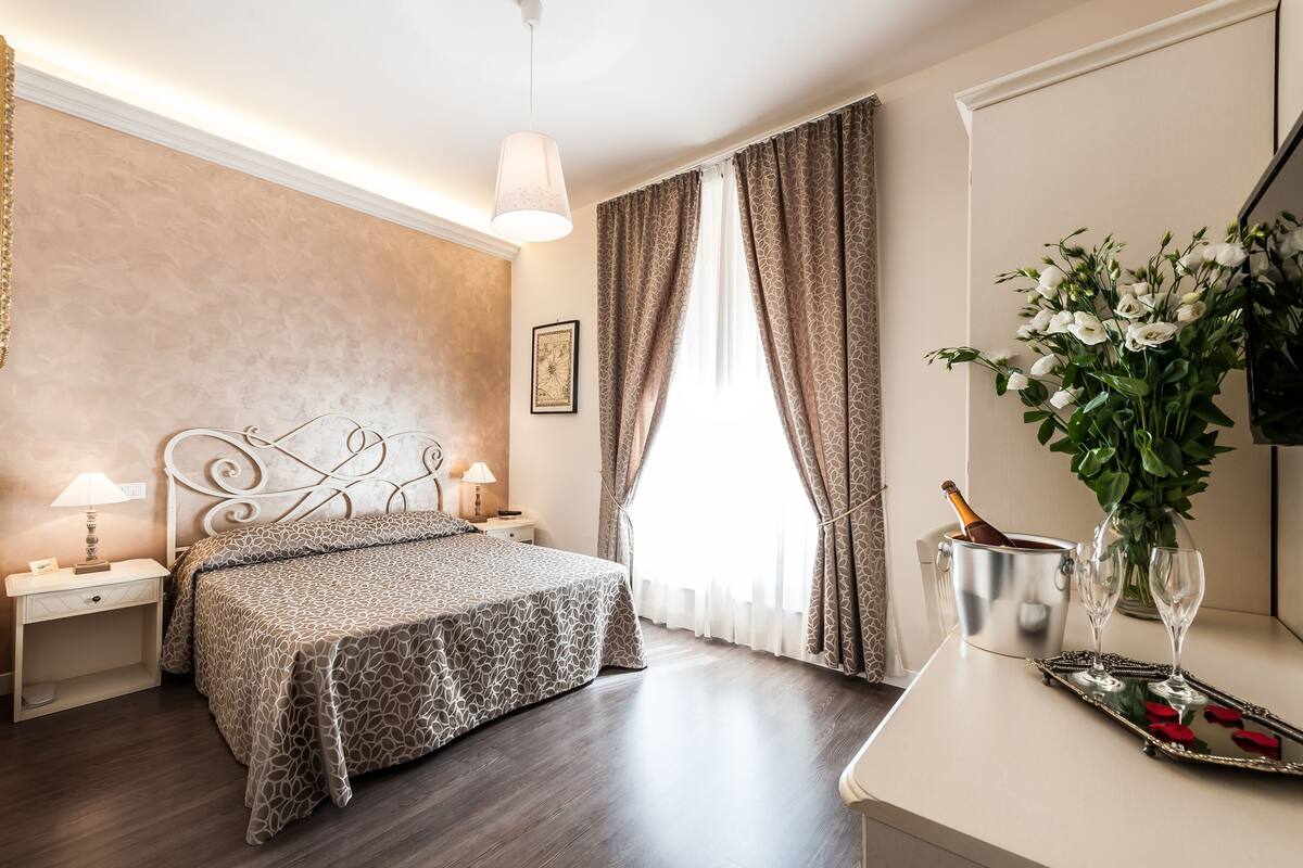 A hotel room with a bed, bedside tables, a nightstand, a chandelier, a window with curtains, a vase with flowers, a tray with rose petals, and a champagne bottle.