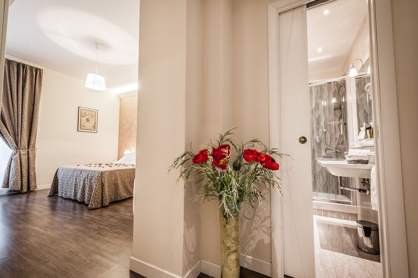 A hotel room with a bed, a vase of red flowers, a bathroom with a glass shower, and a hallway with a door.