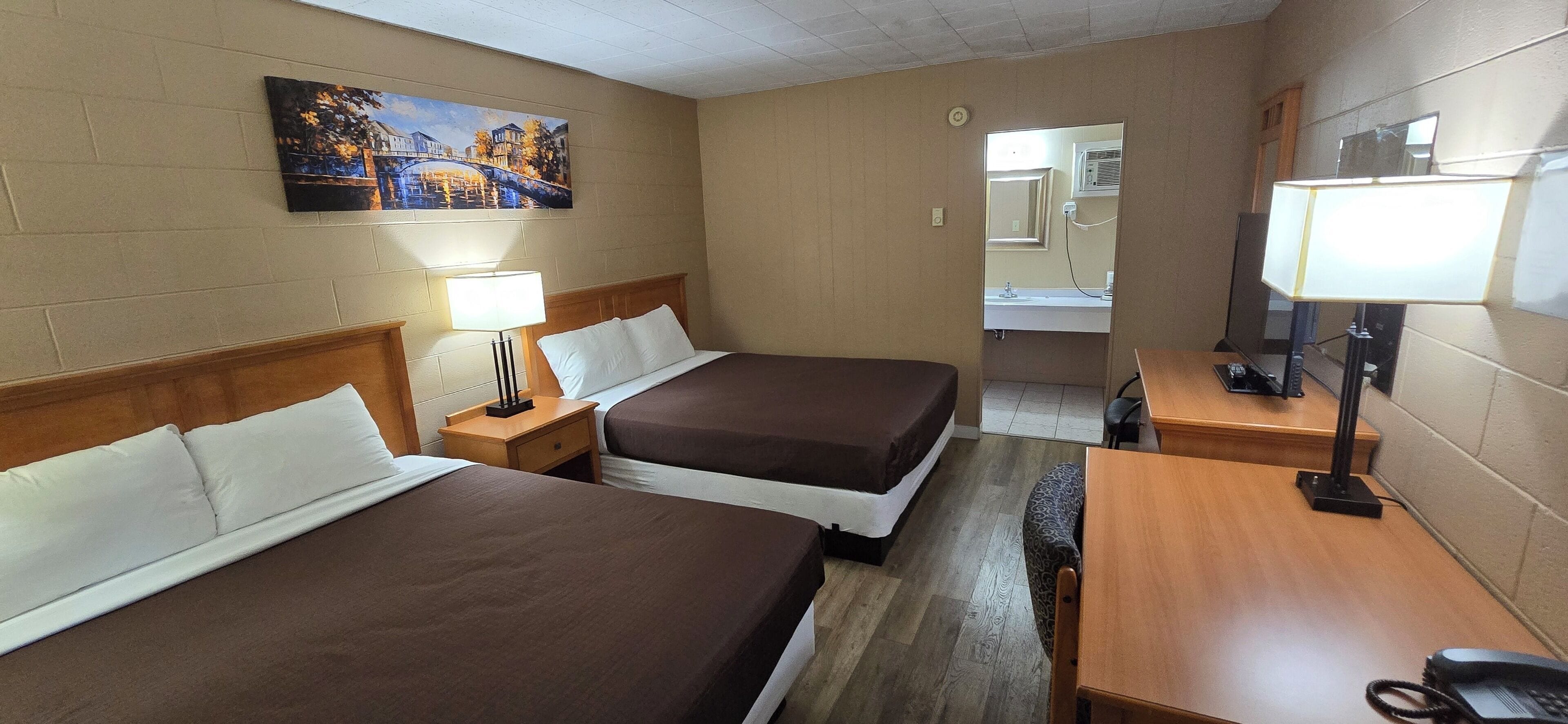 Ein Hotelzimmer mit zwei Betten, einem Schreibtisch, einem Stuhl, einer Lampe, einem Spiegel und einem Bild an der Wand.