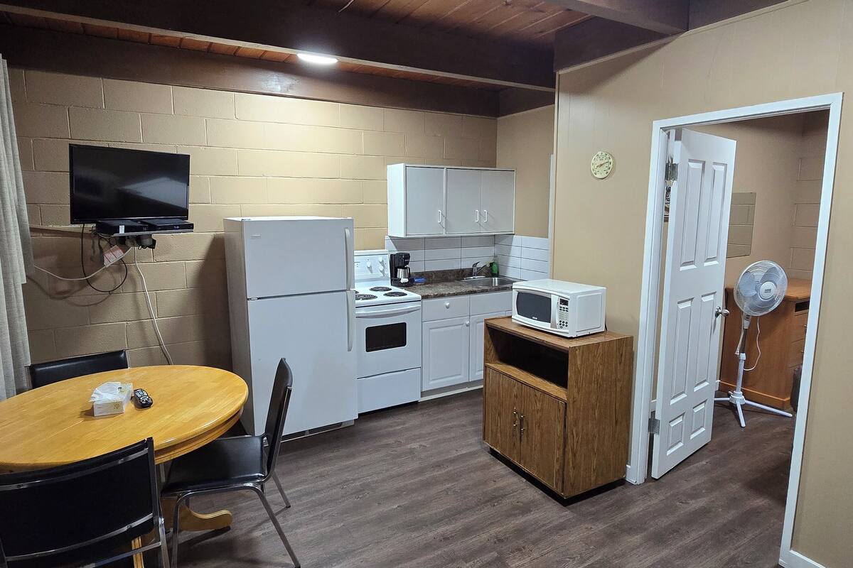 Cocina compacta con refrigerador, microondas y estufa. Hay una mesa de comedor de madera con sillas. Un televisor está montado en la pared y un ventilador cerca de la puerta.
