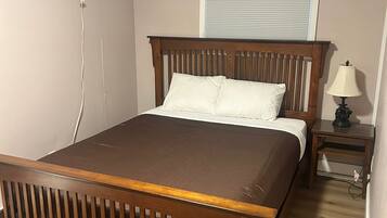 A bedroom with a wooden bed, a bedside table with a lamp, a wall-mounted air conditioner, and a window with blinds.