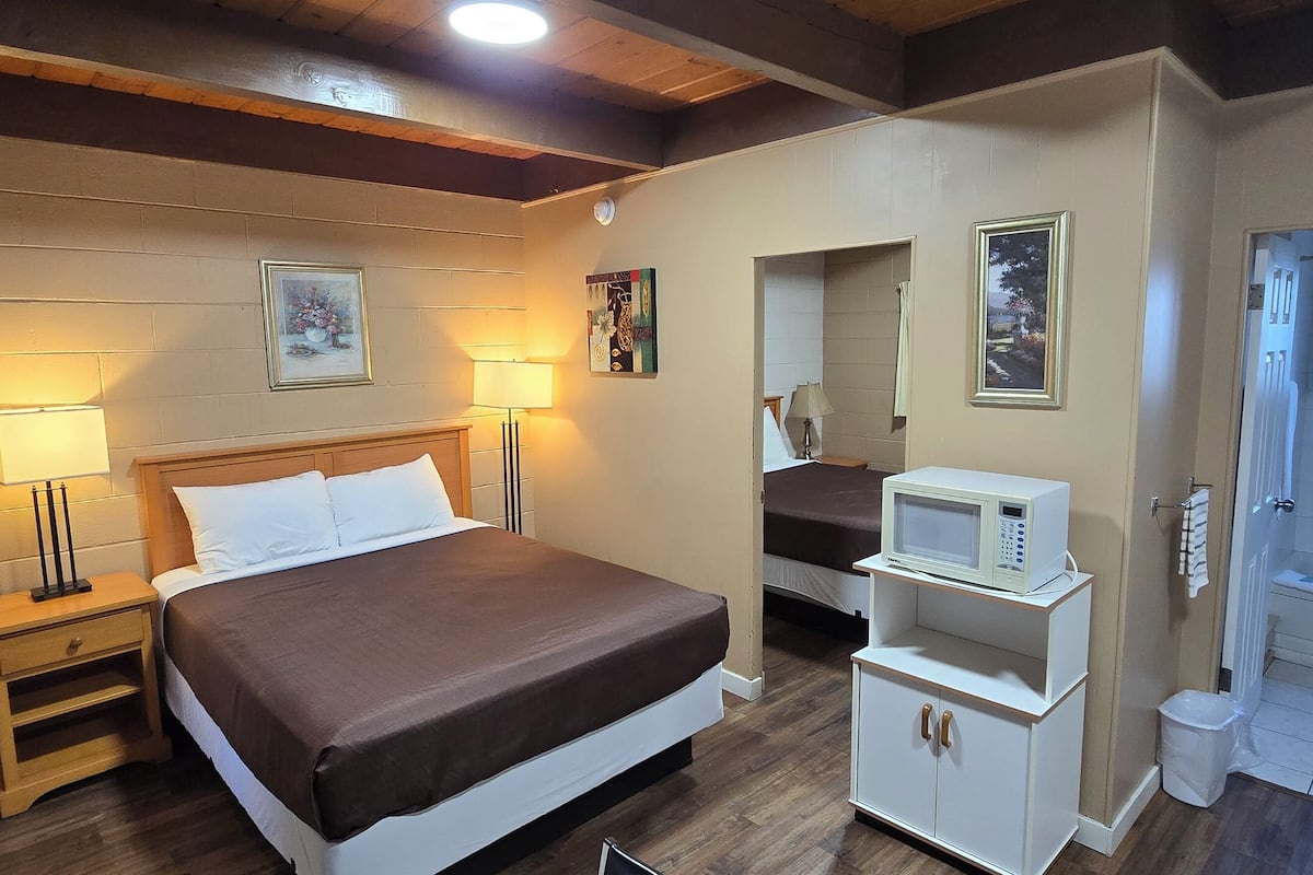 Un dormitorio con cama, lámparas de noche, microondas, un mueble pequeño y un baño con lavamanos y espejo.