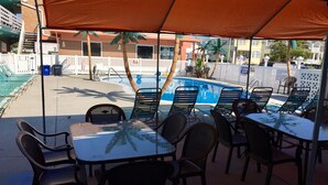 Area piscina all'aperto con sedie a sdraio e tavoli sotto una tettoia, segnale di tuffo e vista sulla piscina e sugli edifici circostanti.