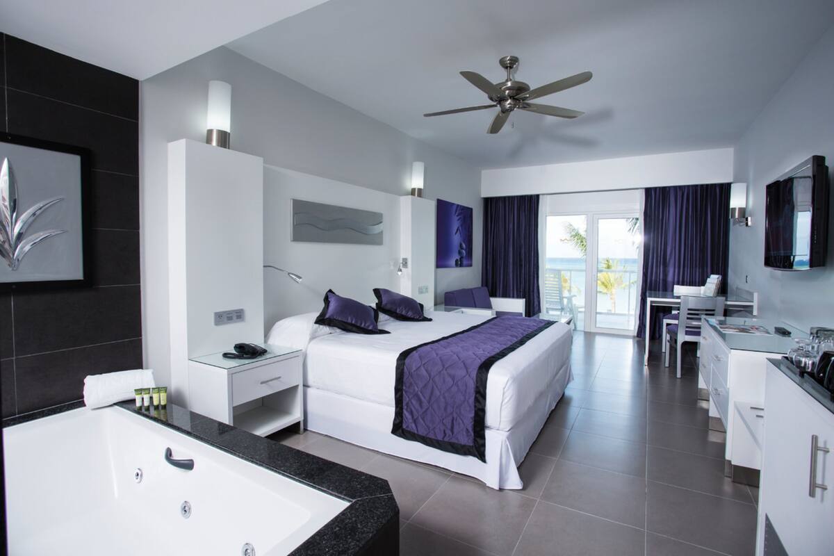 A modern hotel room with a large bed, a separate bathroom, a dining area, and a view of the outdoors.