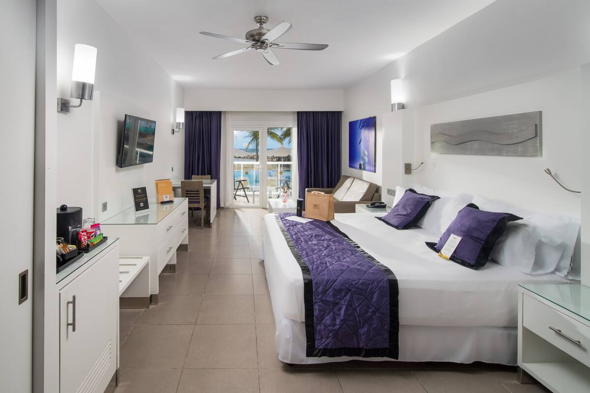 A hotel room with a bed, a desk, a chair, a ceiling fan, and a view of the pool.