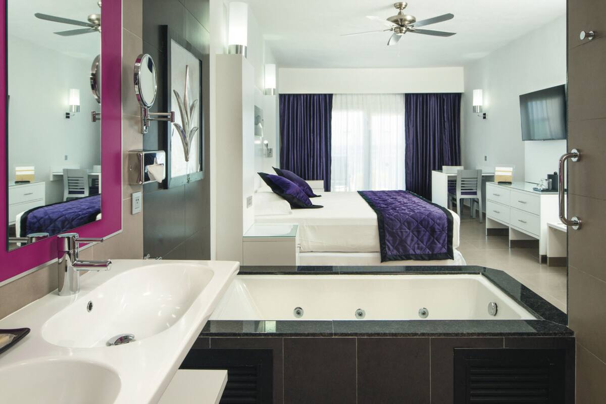 A modern hotel room with a large bathtub, a separate shower area, a bed with a purple blanket, and a desk with a chair.
