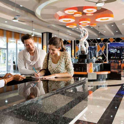 A modern hotel lobby with a reception desk, a large decorative sculpture, and a seating area with orange couches.