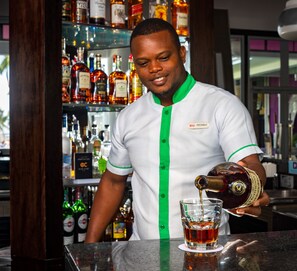 A bartender pouring a drink into a glass at a bar with shelves of liquor bottles in the background.