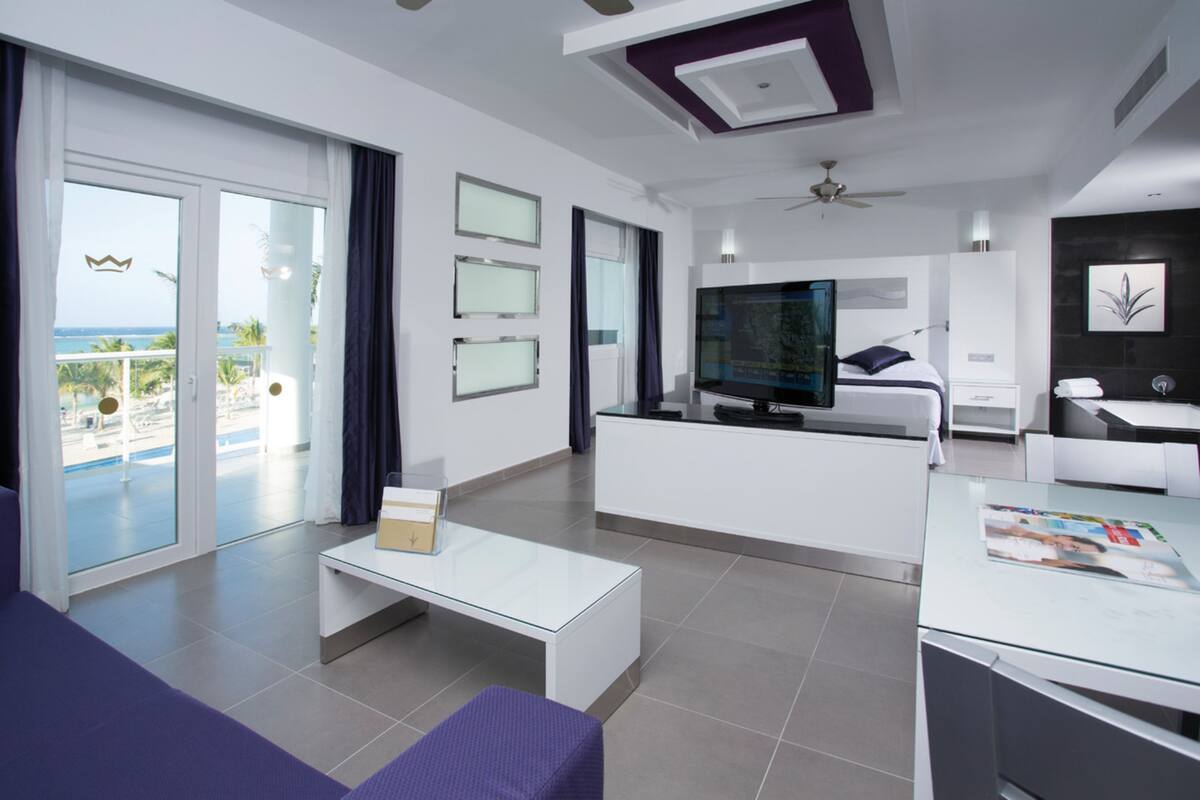 A modern hotel room with a balcony, a flat-screen TV, a sofa, and a dining area.