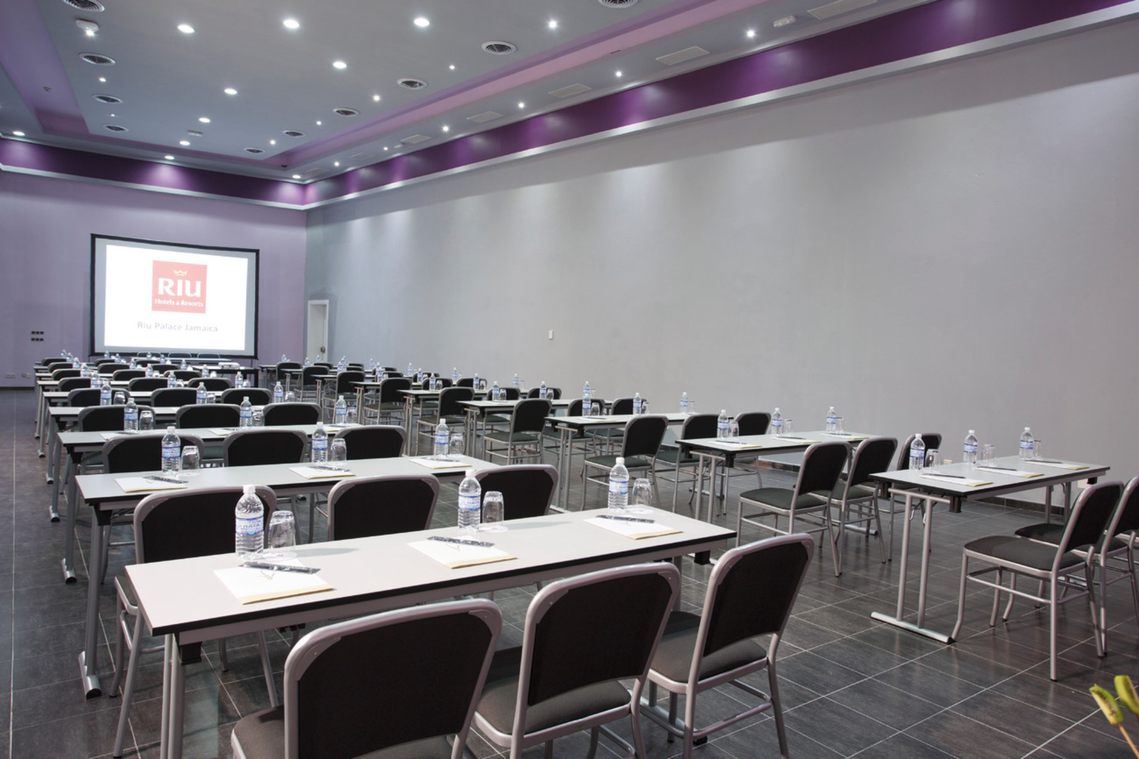 A conference room with multiple tables and chairs, equipped with water bottles and papers, and a projector screen displaying "RIU Hotels & Resorts".