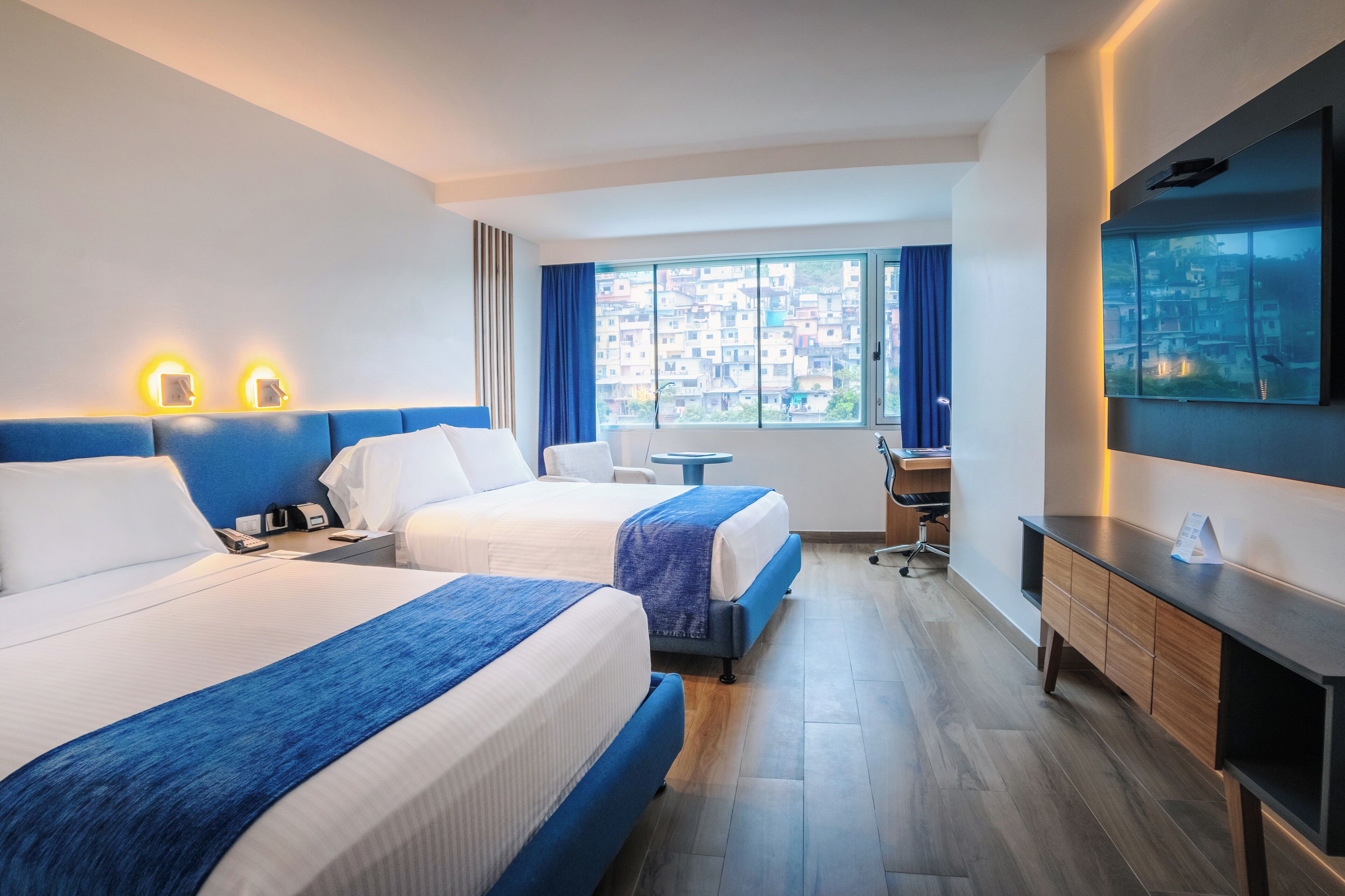 Habitación de hotel con dos camas, un escritorio, una silla, un televisor y una ventana con vistas a la ciudad.