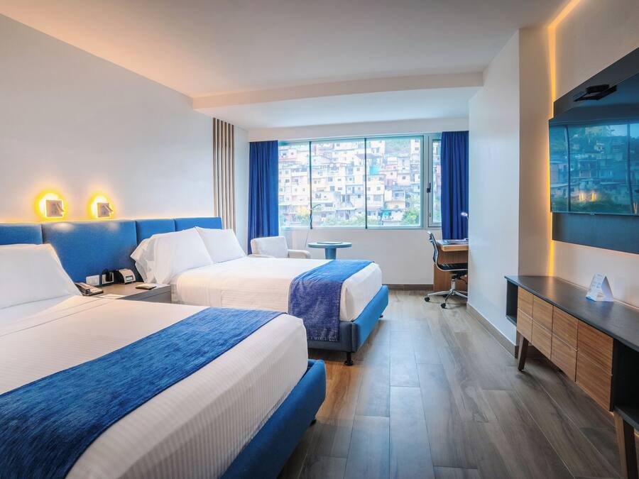 A hotel room with two beds, a desk, a chair, a TV, and a window with a city view.