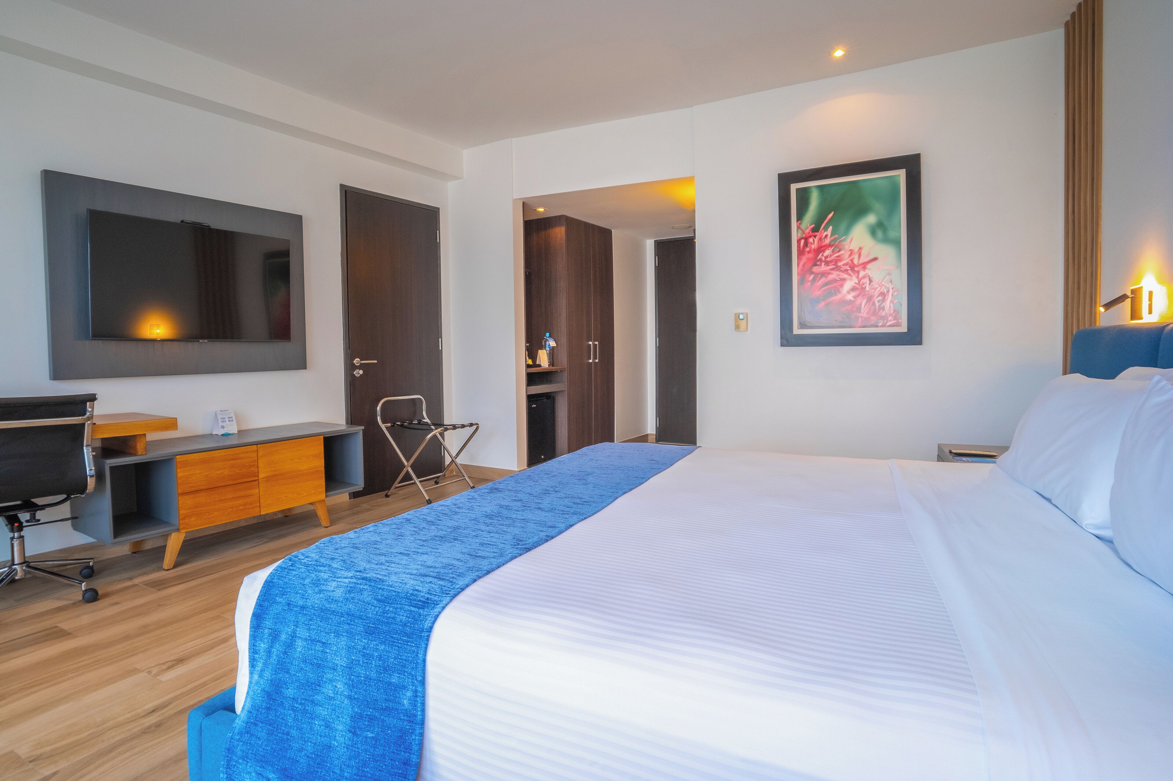 A modern hotel room with a large bed, a desk, a chair, and a wardrobe.