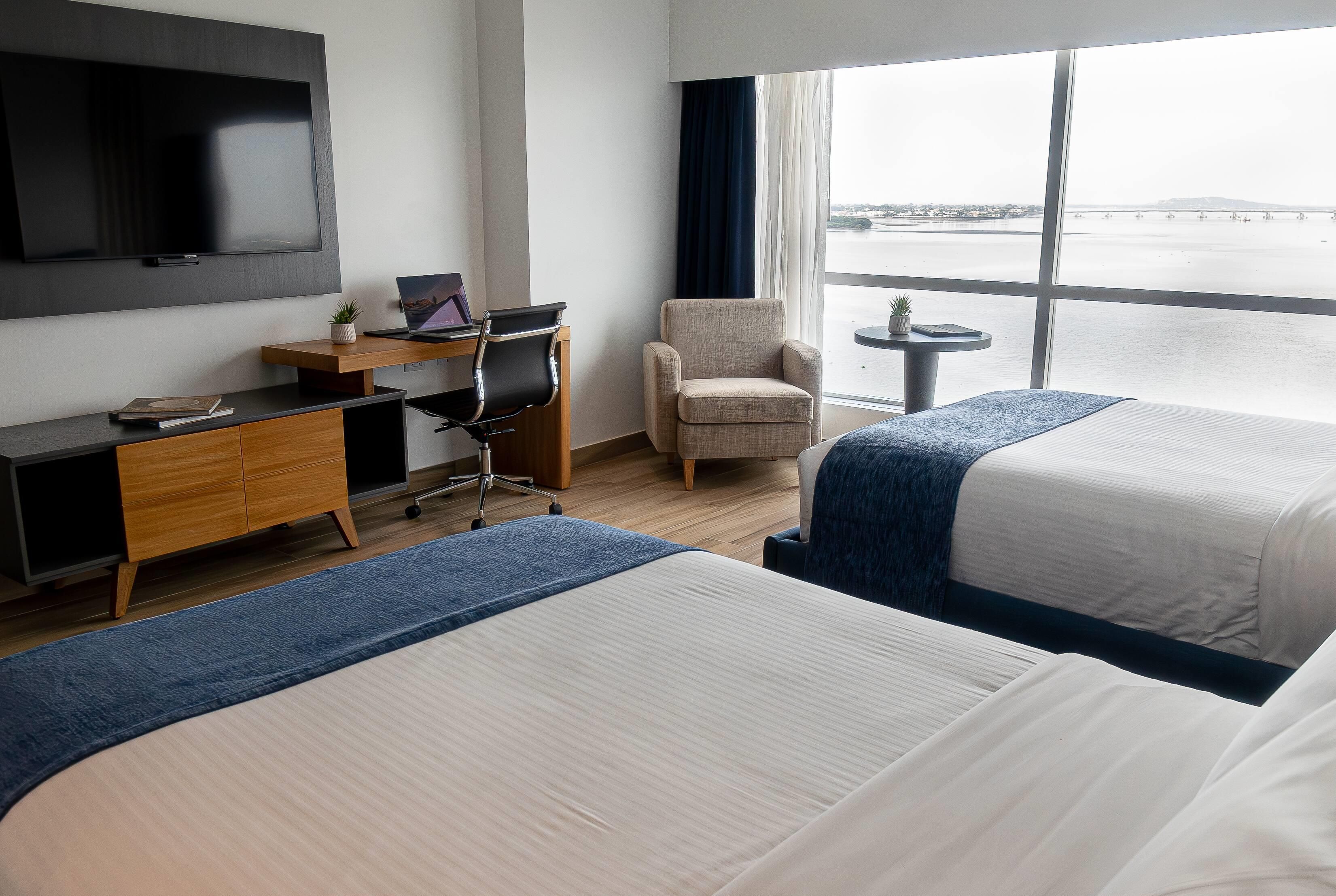 A hotel room with a bed, a desk with a laptop, a chair, a TV, and a window with a view of a body of water.
