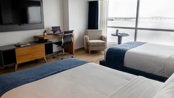 A hotel room with a bed, a desk with a laptop, a chair, a TV, and a window with a view of a body of water.
