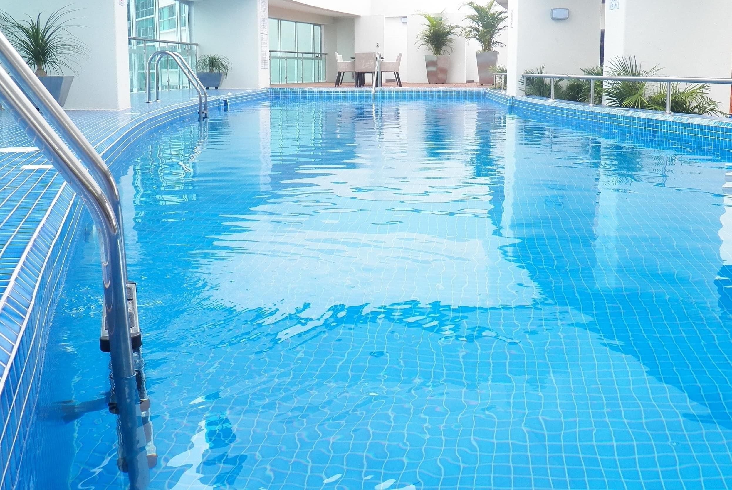 A clear indoor swimming pool with a metal ladder, surrounded by potted plants and a modern building facade.