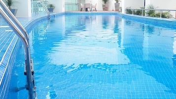 A clear indoor swimming pool with a metal ladder, surrounded by potted plants and a modern building facade.