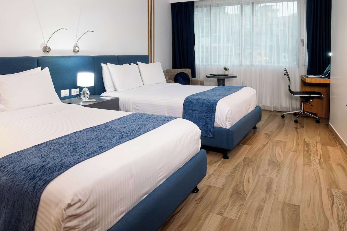 Habitación de hotel con dos camas, un escritorio, una silla, una mesita y una ventana con cortinas.