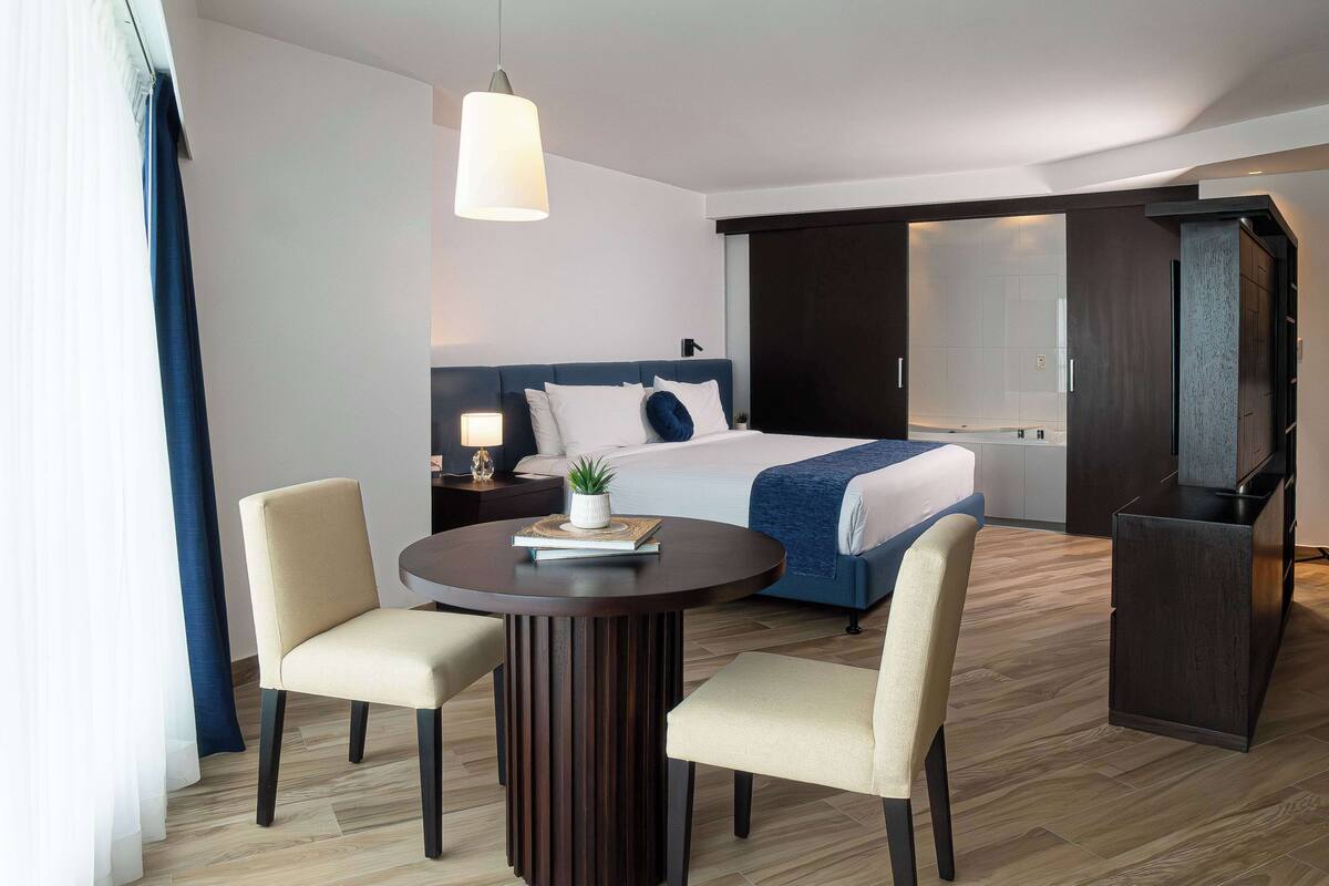 Una habitación de hotel moderna con una cama grande, una mesa redonda de comedor con dos sillas, una mesita auxiliar con una lámpara y un espejo grande.