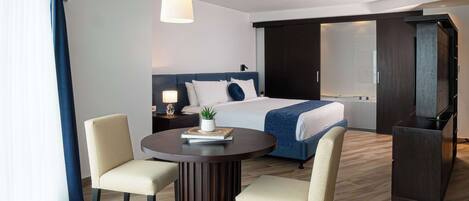 A modern hotel room with a large bed, a round dining table with two chairs, a small side table with a lamp, and a large mirror.