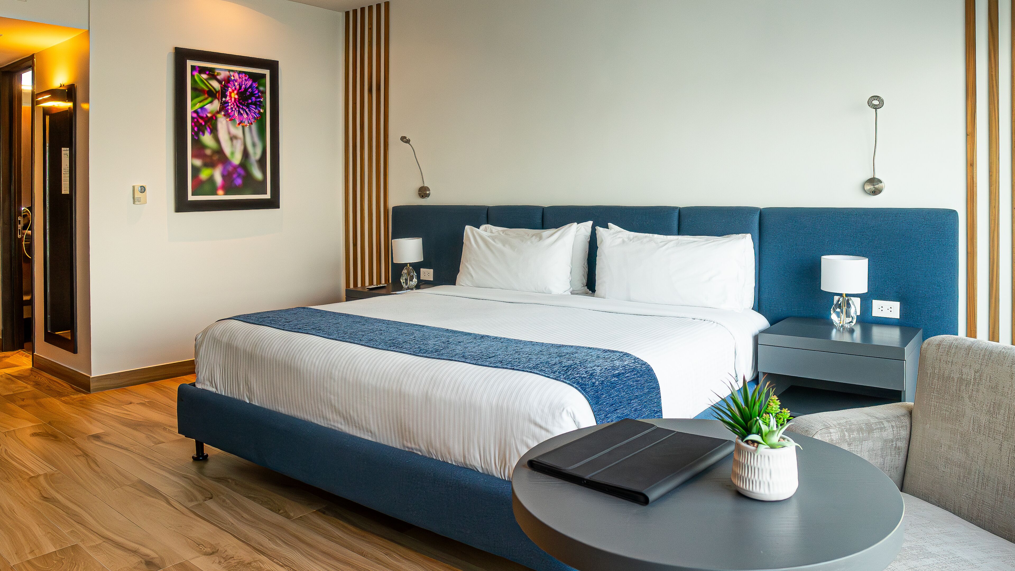 Une chambre d’hôtel moderne avec un grand lit, une table de chevet avec une lampe, une petite table d’appoint, une table ronde avec une tablette et une plante, et un plancher en bois.