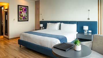 A modern hotel room with a large bed, a bedside table with a lamp, a small side table, a round table with a tablet and a plant, and a wooden floor.