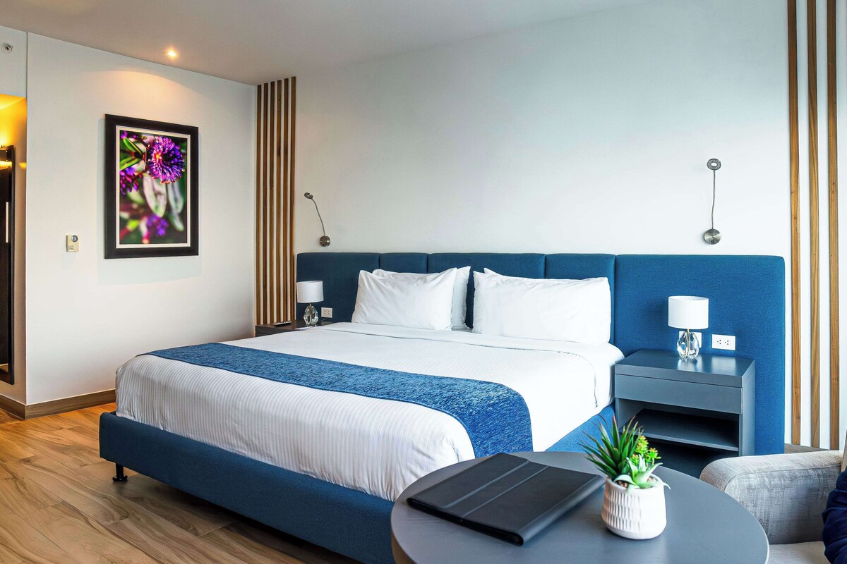 A modern hotel room with a large bed, a bedside table, a lamp, a small plant, and a laptop on a round table.