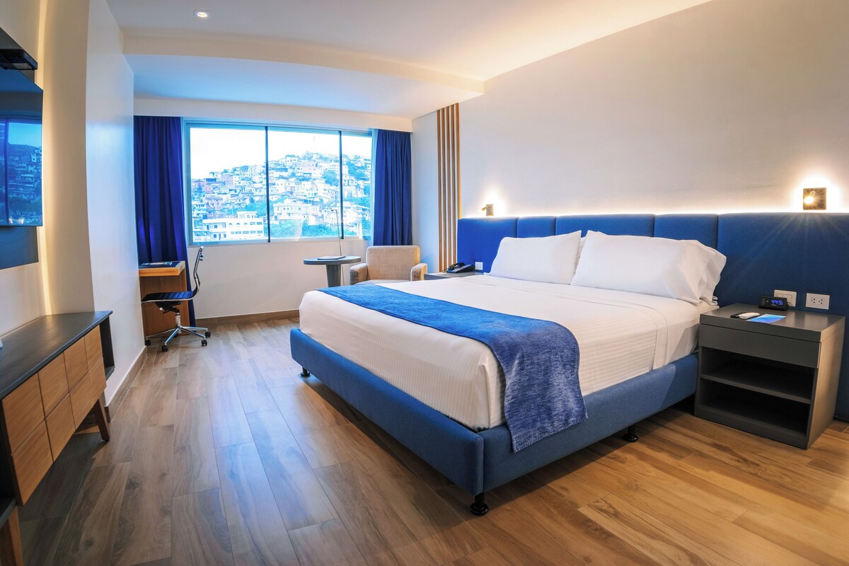 Una habitación de hotel moderna con una cama grande, un escritorio, una silla y vistas a un paisaje urbano a través de la ventana.
