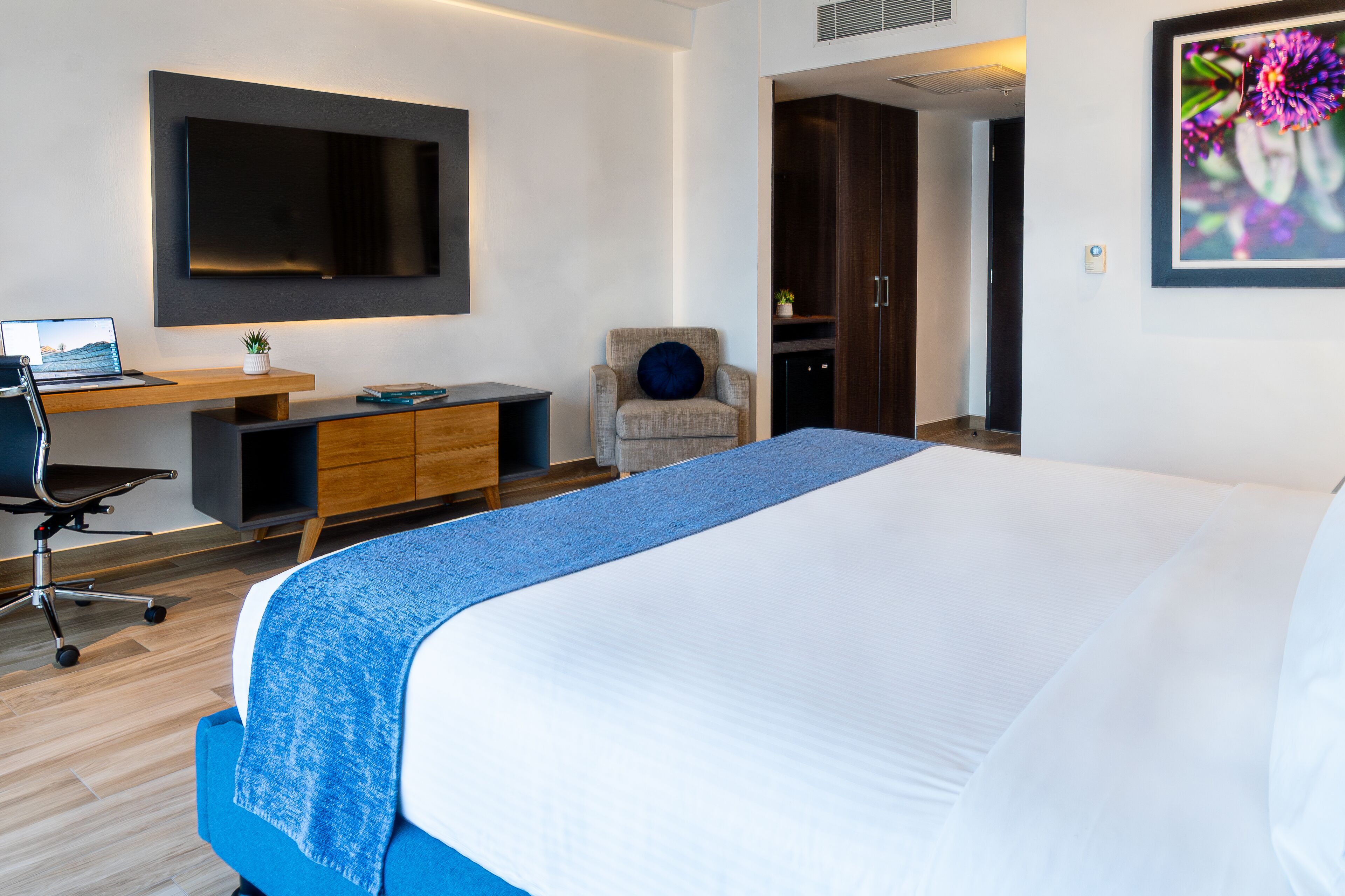 Una habitación de hotel moderna con una cama grande, un escritorio con una computadora portátil, una silla y un televisor montado en la pared.