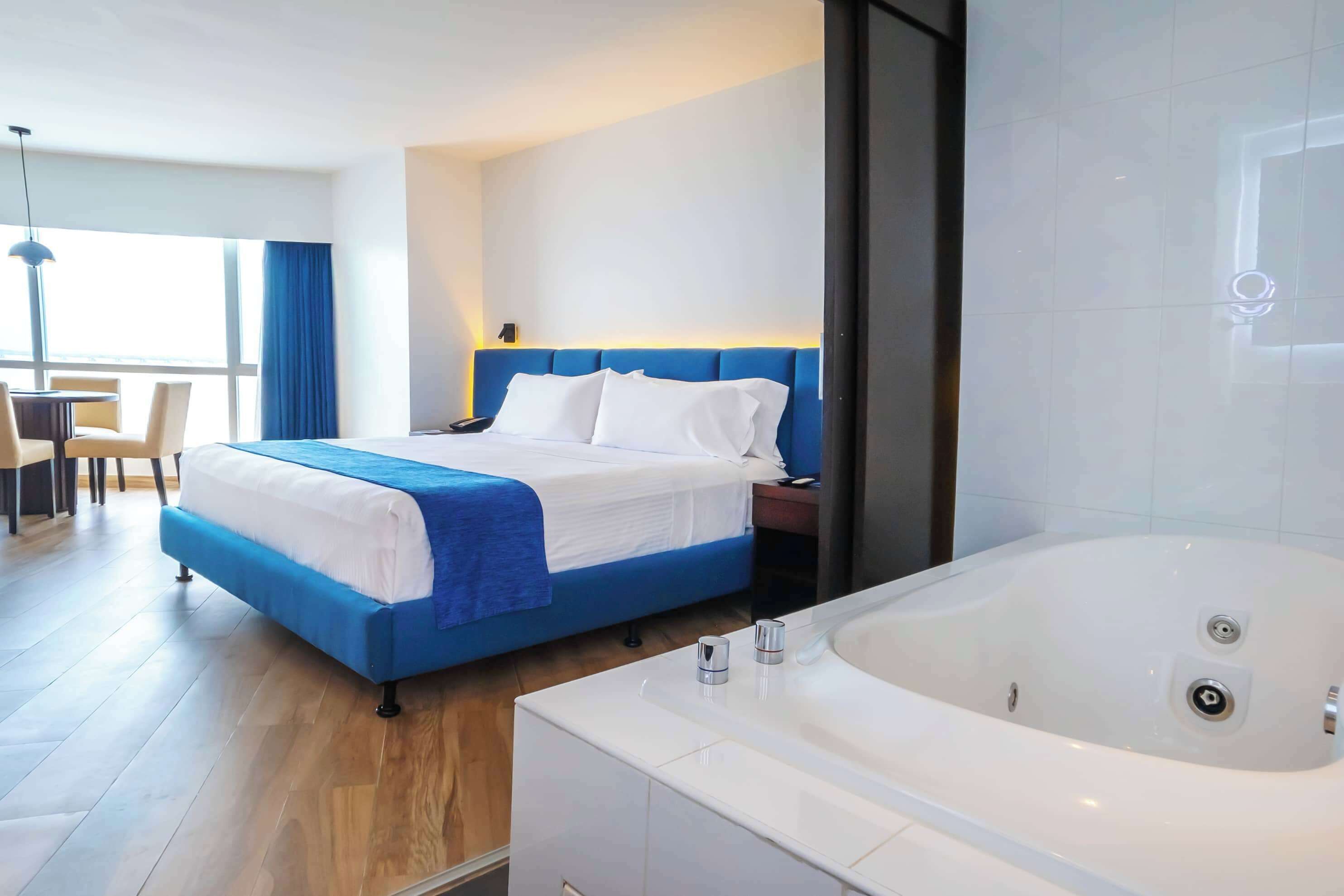 Una habitación de hotel moderna con una cama grande, un baño independiente con jacuzzi y un comedor con mesa y sillas.