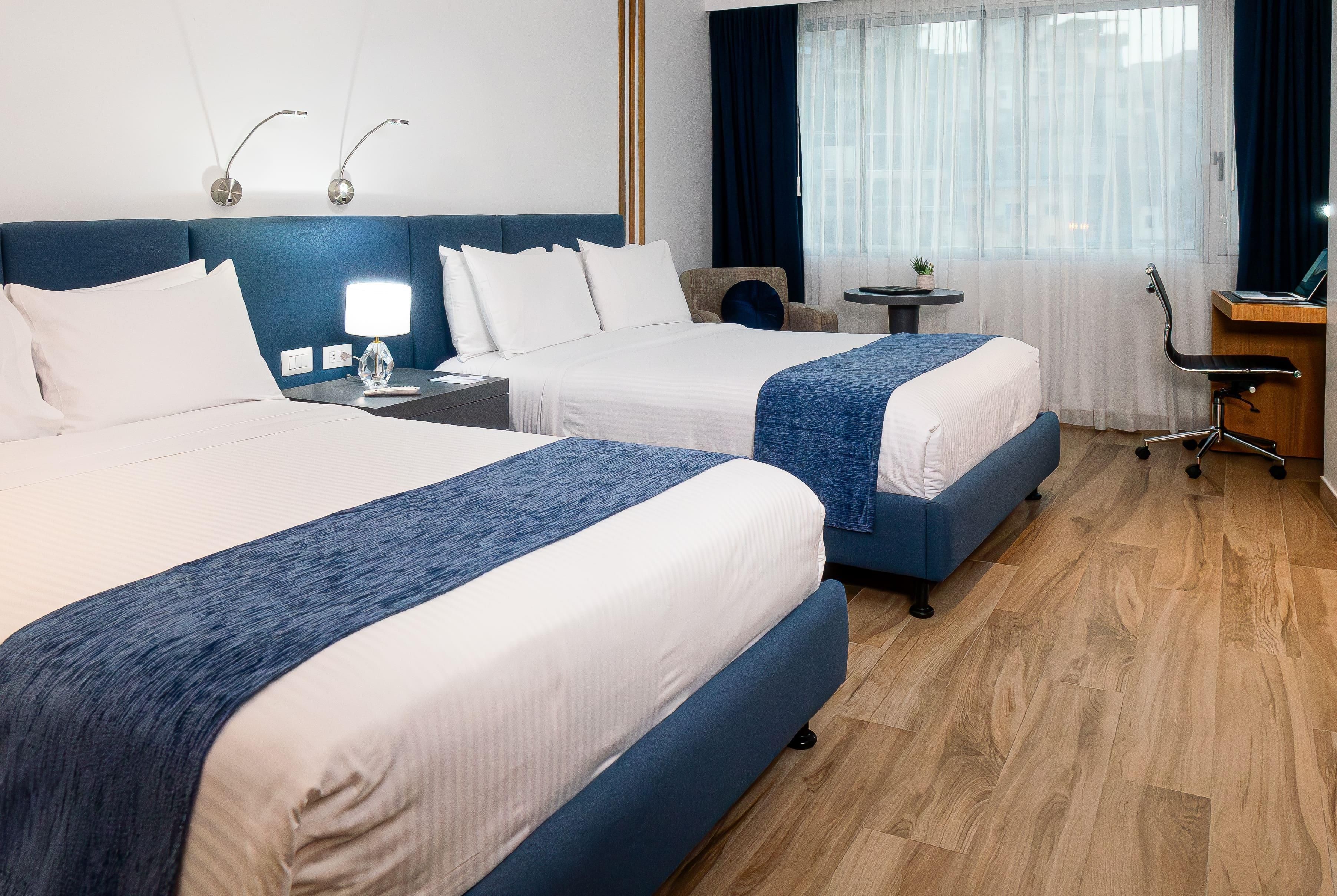 A hotel room with two beds, a desk, a chair, and a small table.