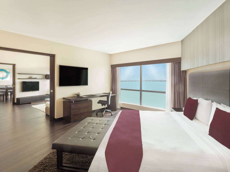 A modern hotel room with a large bed, a desk with a chair, a television, and a view of the sea.