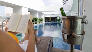 Une personne est assise au bord d’une piscine, un livre à la main et un verre dans un seau en métal.