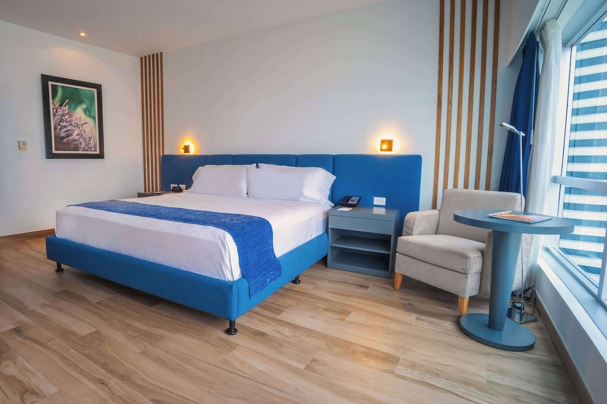 A modern hotel room with a large bed, a desk, a chair, and a window with blinds.
