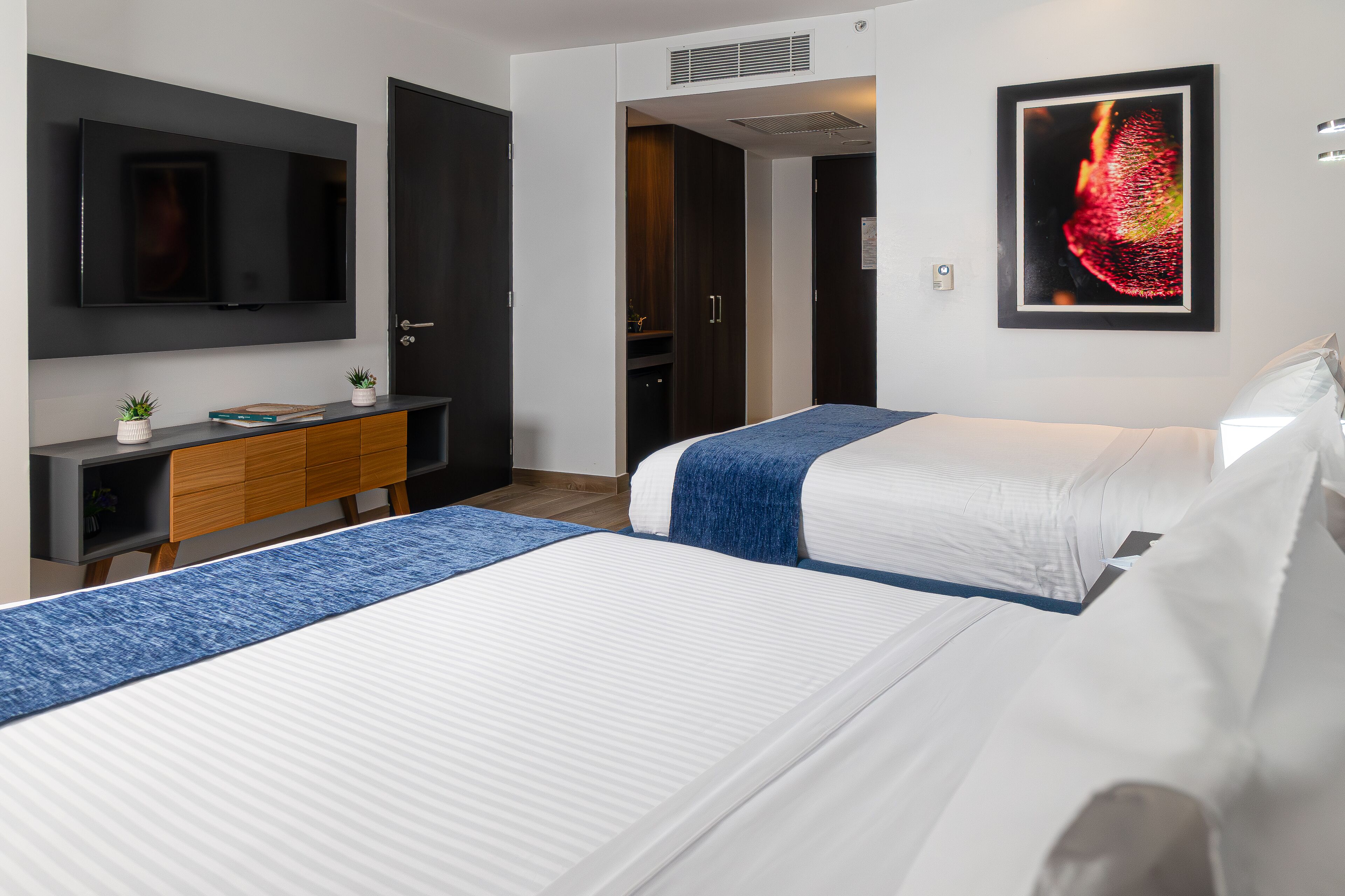 A hotel room with two beds, a TV mounted on the wall, a desk with a computer, and a wardrobe.