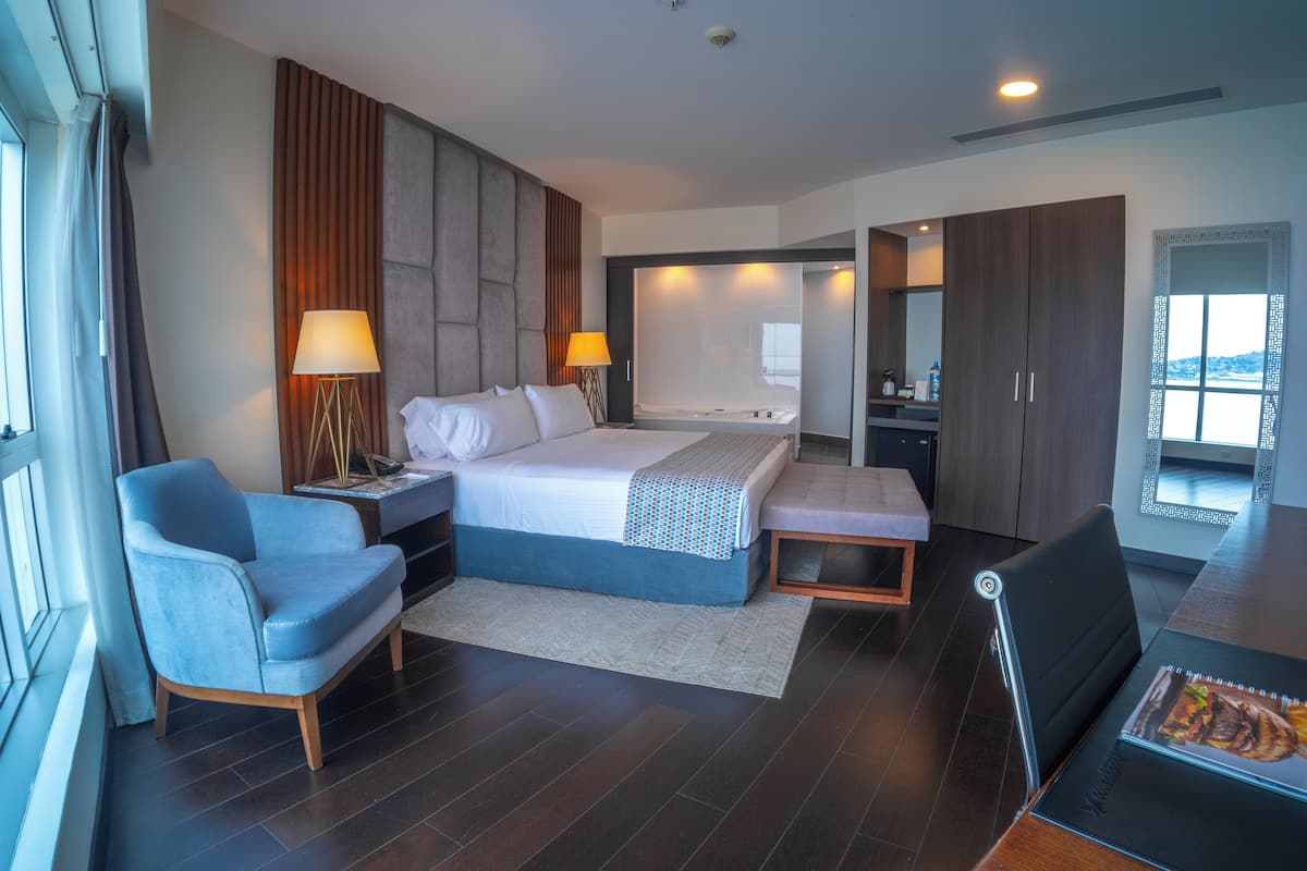 Una habitación de hotel moderna con una cama grande, dos mesitas de noche con lámparas, una zona de estar con una silla y un banco, una mesa de comedor y vistas al exterior a través de una ventana.