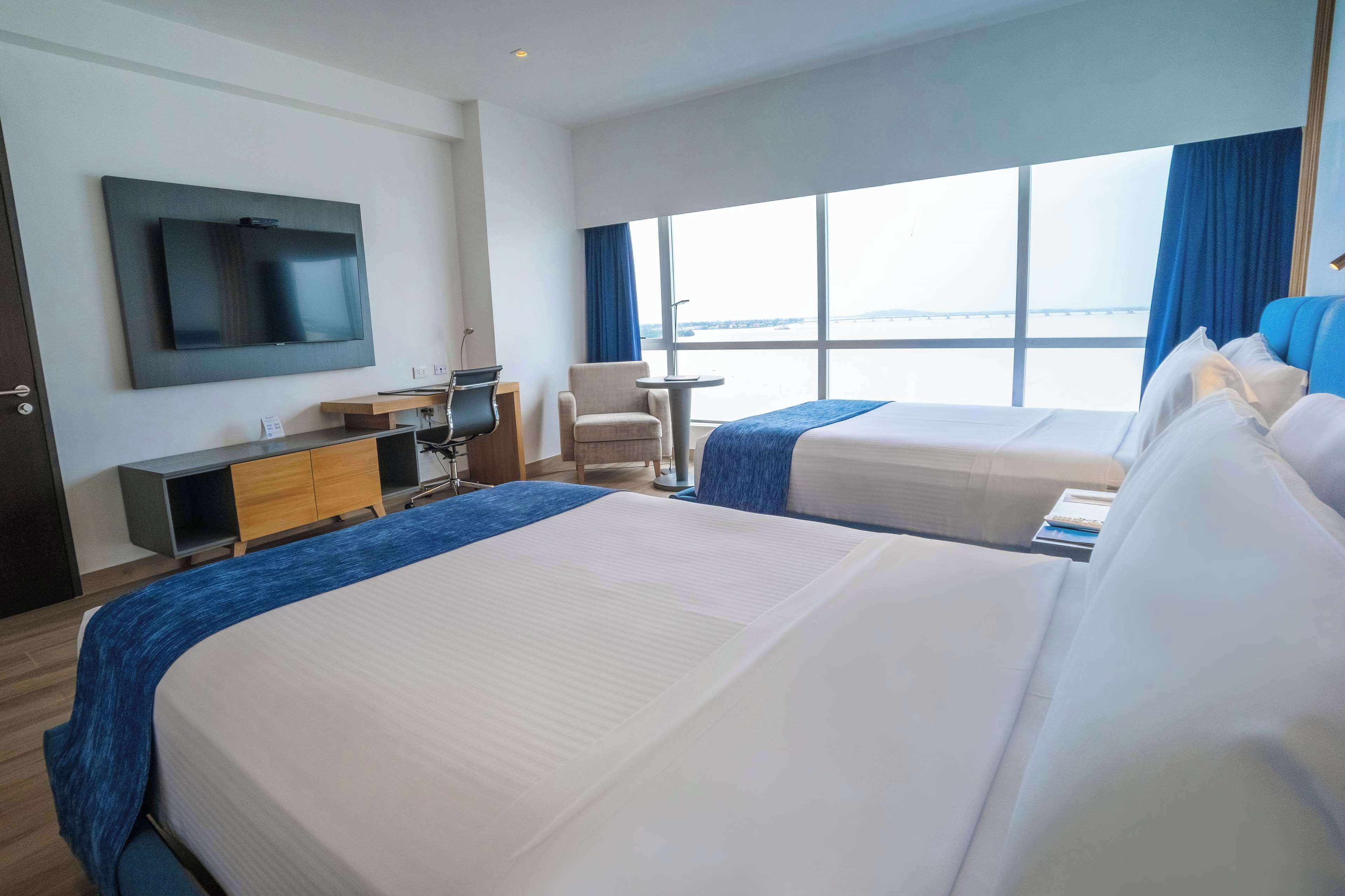 Quarto de hotel com duas camas, uma TV fixada na parede, uma escrivaninha com cadeira e uma janela grande com vista.