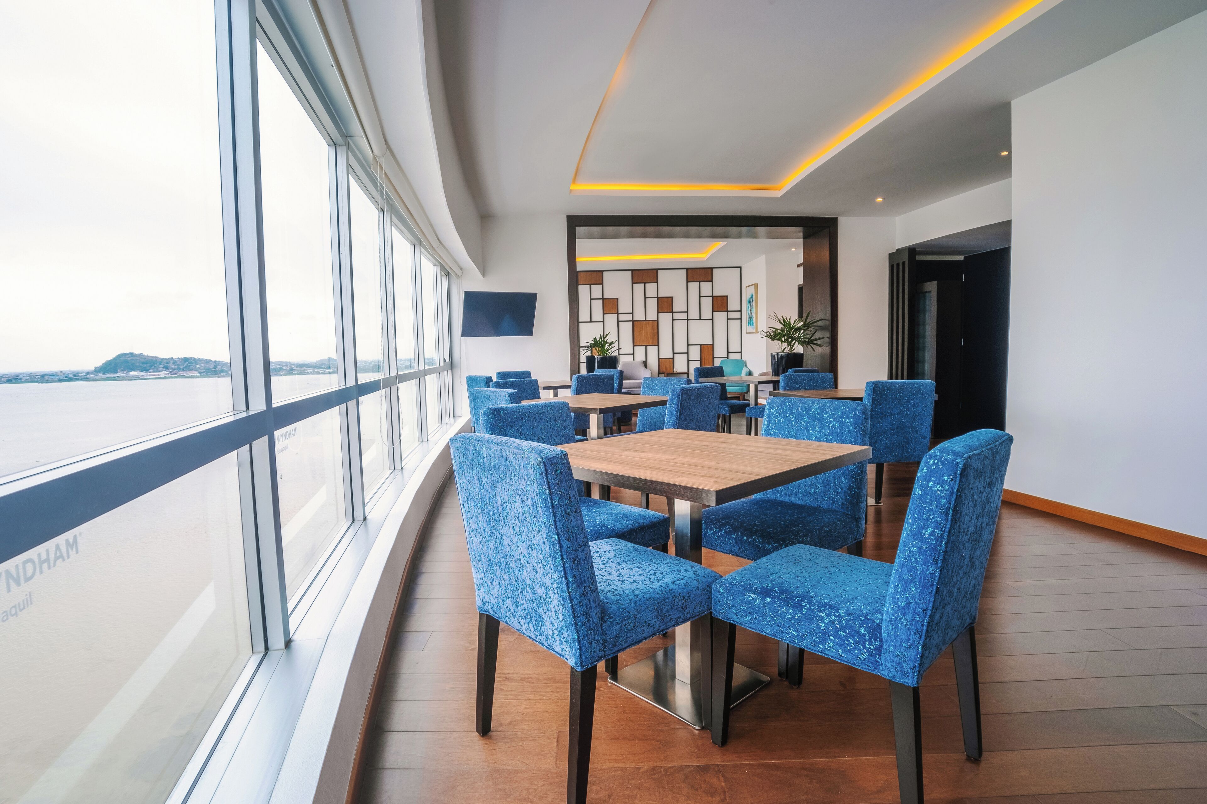 A modern dining area with blue upholstered chairs and a wooden table, large windows with a view of the water, and a contemporary wall design.