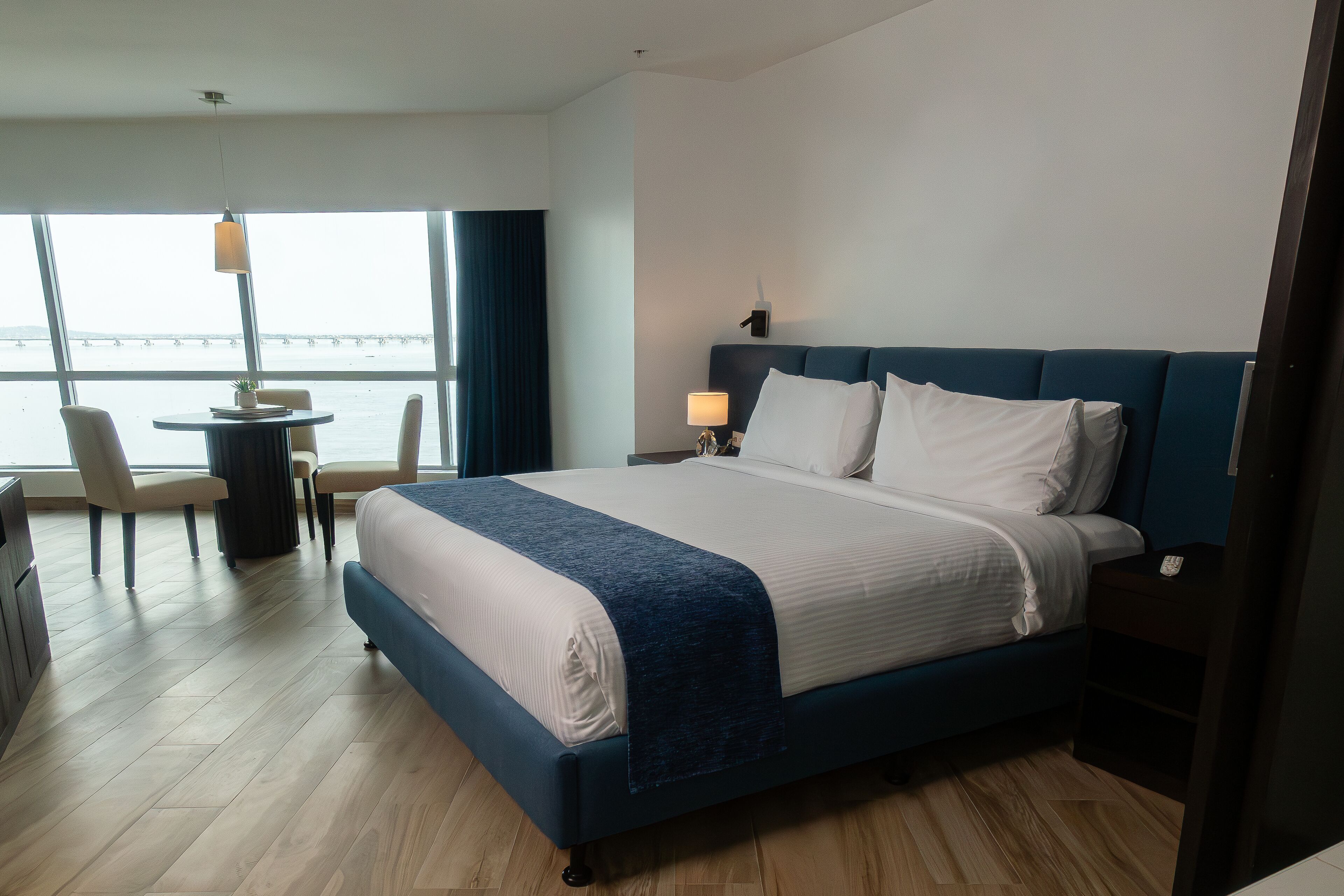 A hotel room with a large bed, a desk with chairs, a lamp, and a view of the sea.