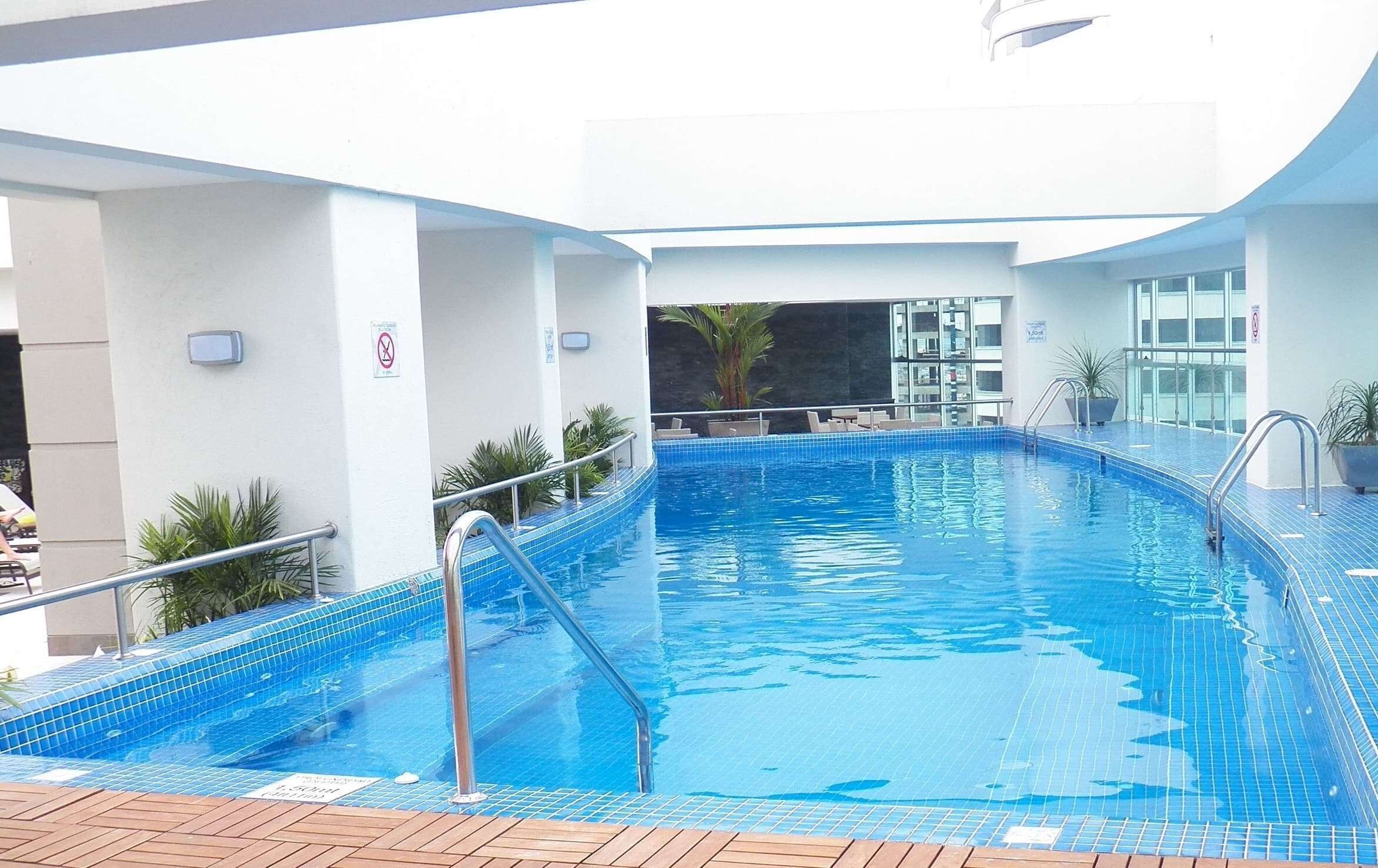 Una piscina interior con suelo de baldosas azules, barandillas de metal y vistas del interior del edificio.