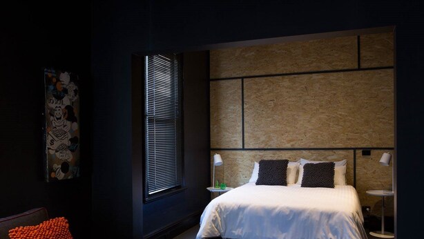 A modern bedroom with a large bed, a side table, a chair, and a window with blinds. - The Franklin Hotel (Adelaide)