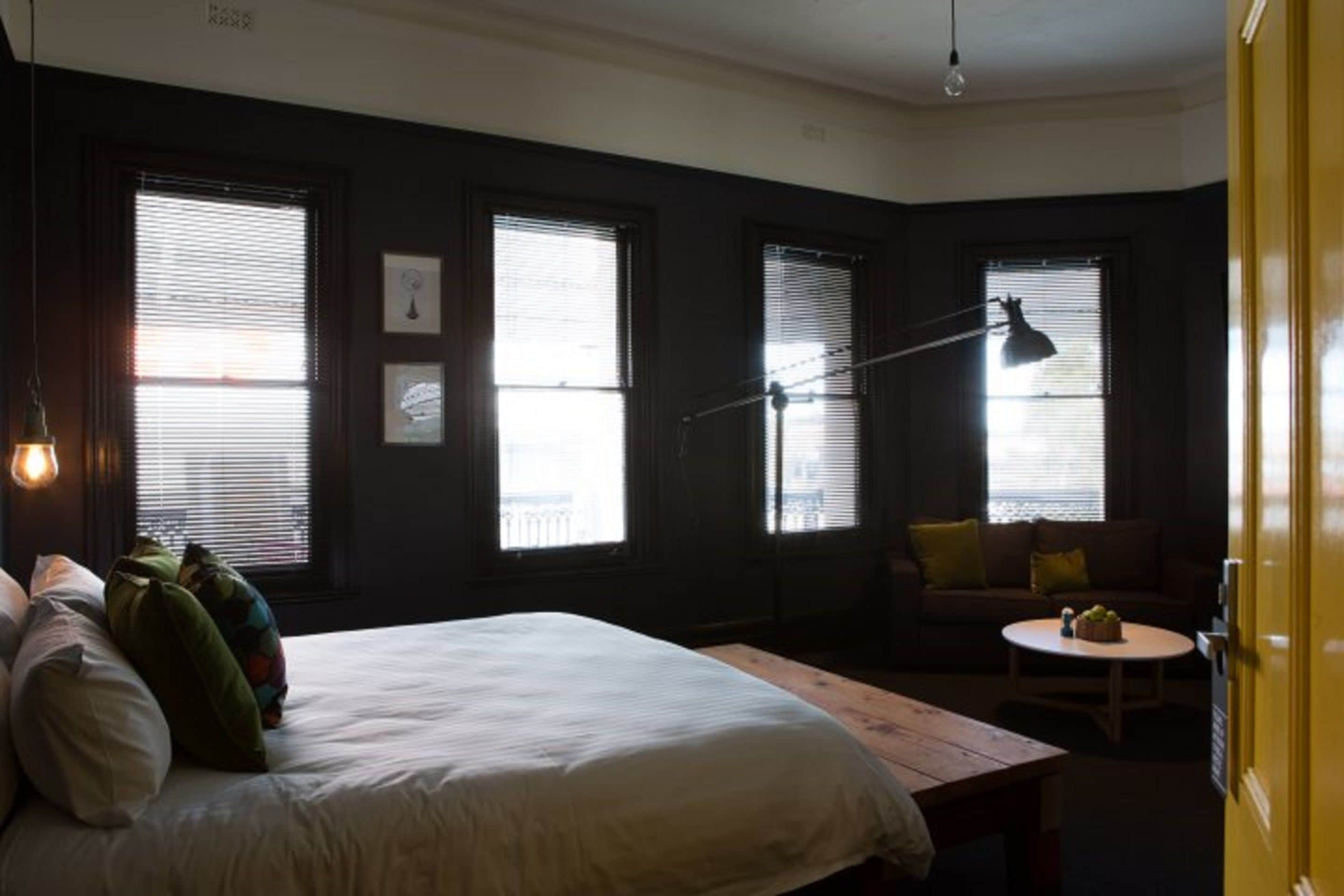 Ein Schlafzimmer mit einem Bett, einem Schreibtisch, einem Sessel und einem Sofa. Es gibt drei Fenster mit Jalousien, eine Lampe und eine Tür.
