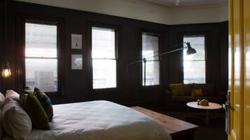 Ein Schlafzimmer mit einem Bett, einem Schreibtisch, einem Sessel und einem Sofa. Es gibt drei Fenster mit Jalousien, eine Lampe und eine Tür.