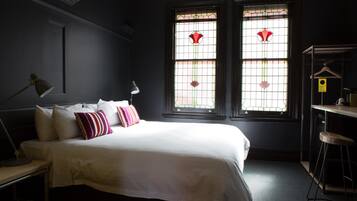 Ein Schlafzimmer mit einem großen Bett, zwei Nachttischlampen, einem Schreibtisch mit Stuhl und zwei Fenstern mit verzierten Glasscheiben.