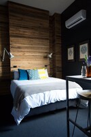 Une chambre moderne avec un mur d’accent en bois, un lit avec une literie blanche et des coussins colorés, un bureau avec un vase et un appareil de climatisation.