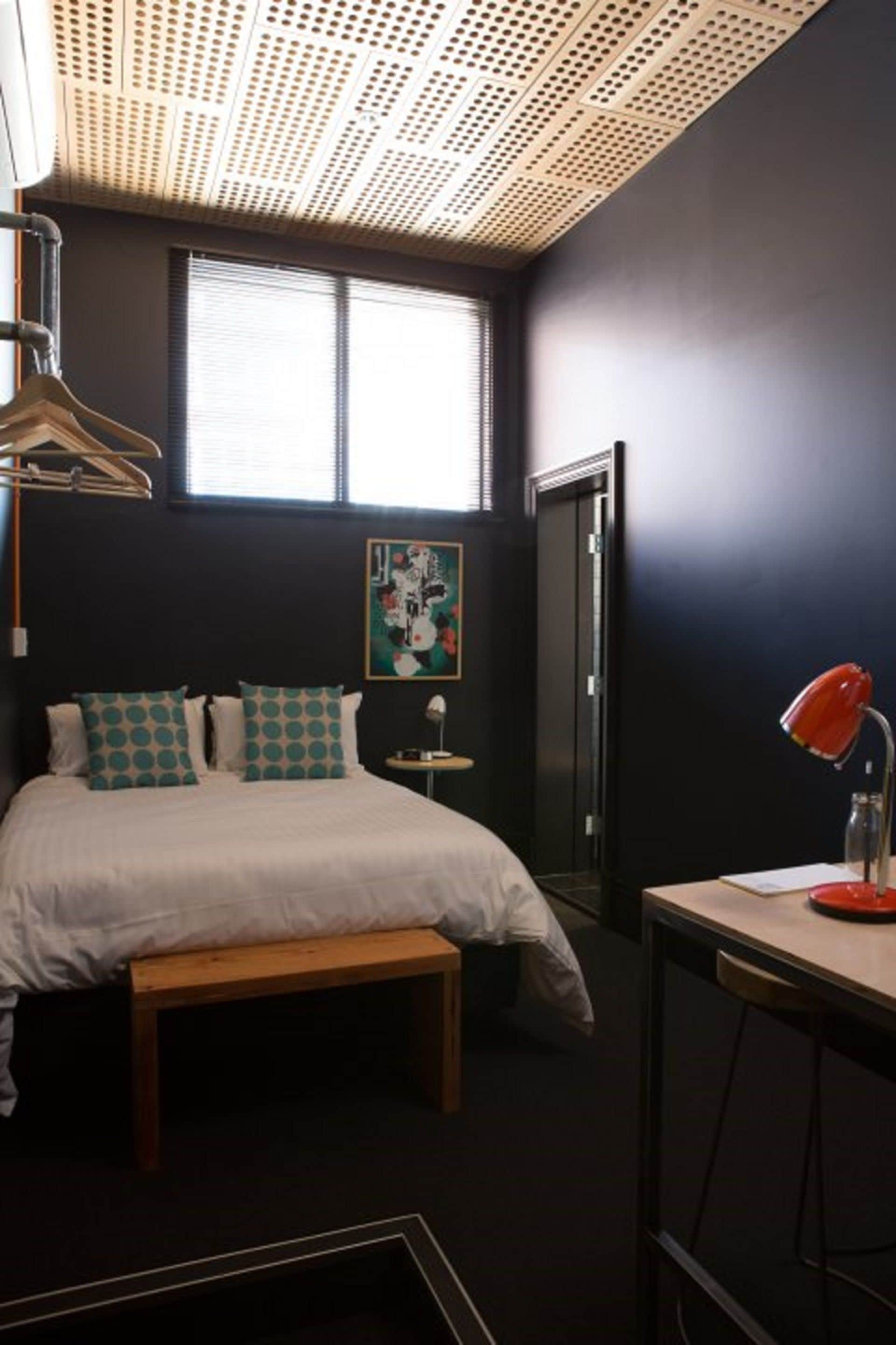 A bedroom with a bed, a bench, a desk with a lamp, a painting on the wall, and a window with blinds.