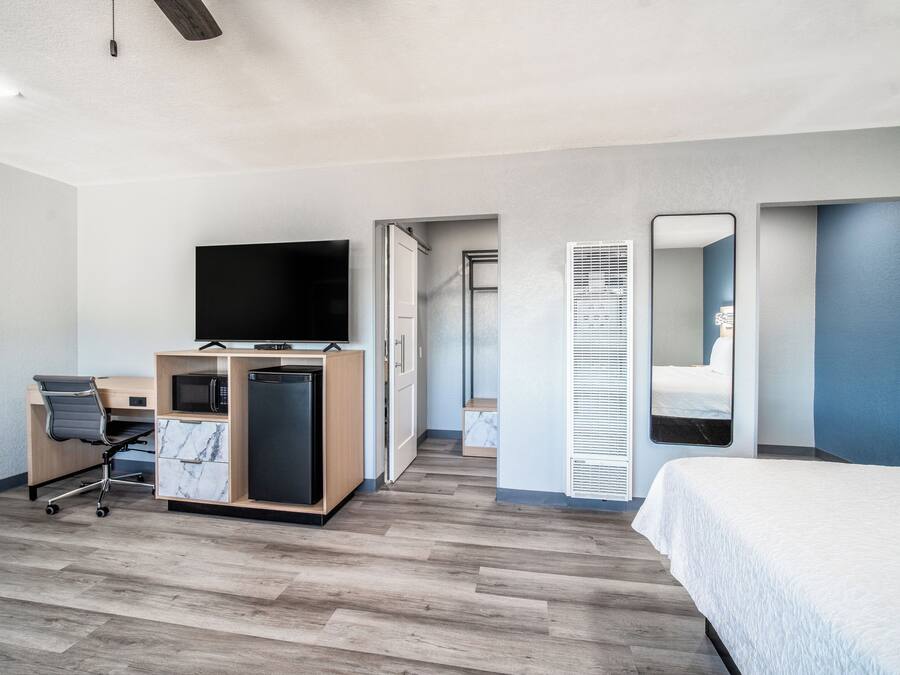 Una habitación de hotel moderna con suelo de madera, una cama, un escritorio con silla, un televisor y un baño con espejo e inodoro.