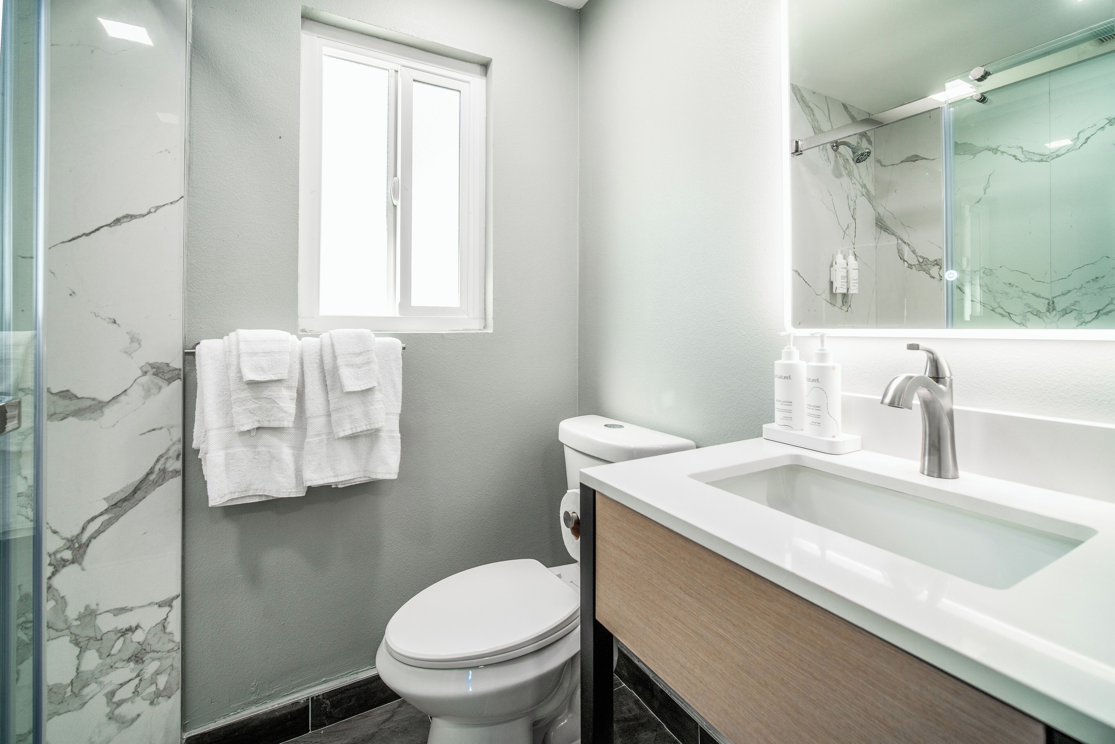 A modern bathroom with a marble wall, a toilet, a sink with a mirror, and a window.